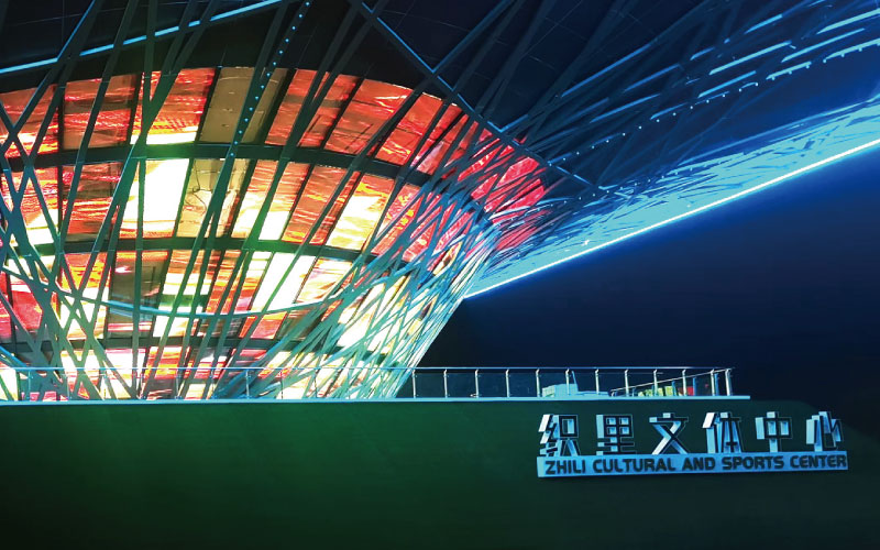 位（wèi）於浙江湖州的（de）織裏（lǐ）文體中（zhōng）心，是一座大型異形鋼架結構建築。色多多AV导航為其定製近1000平米透明屏，運用（yòng）高清LED透明屏及控製係統，將其打造成了（le）一幅巨型建築媒體立麵。